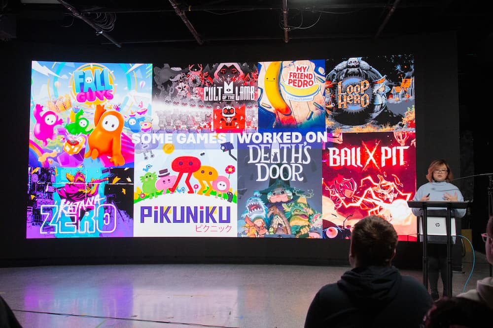 Clara Sia presents to a crowd, her video game career background on display. There are games like Ball X Pitt, PikuNiku, and Gall Guys to name a few.